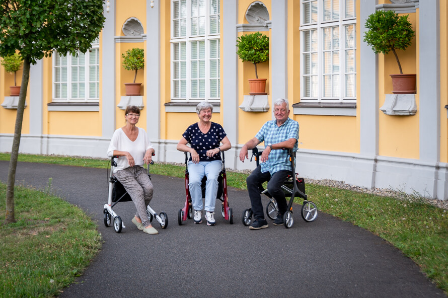 Drei Senioren sitzen im Halbkreis auf ihren Rollatoren im Park.