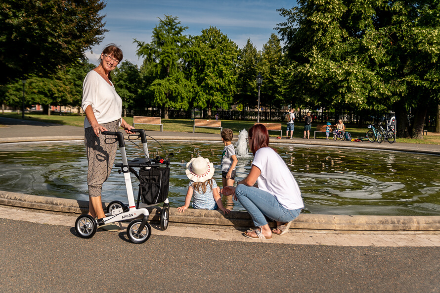 Eine ältere Frau steht mit ihrem Rollator an einem Springbrunnen. Rechts daneben hockt eine Frau, deren zwei Kinder im Wasser spielen.